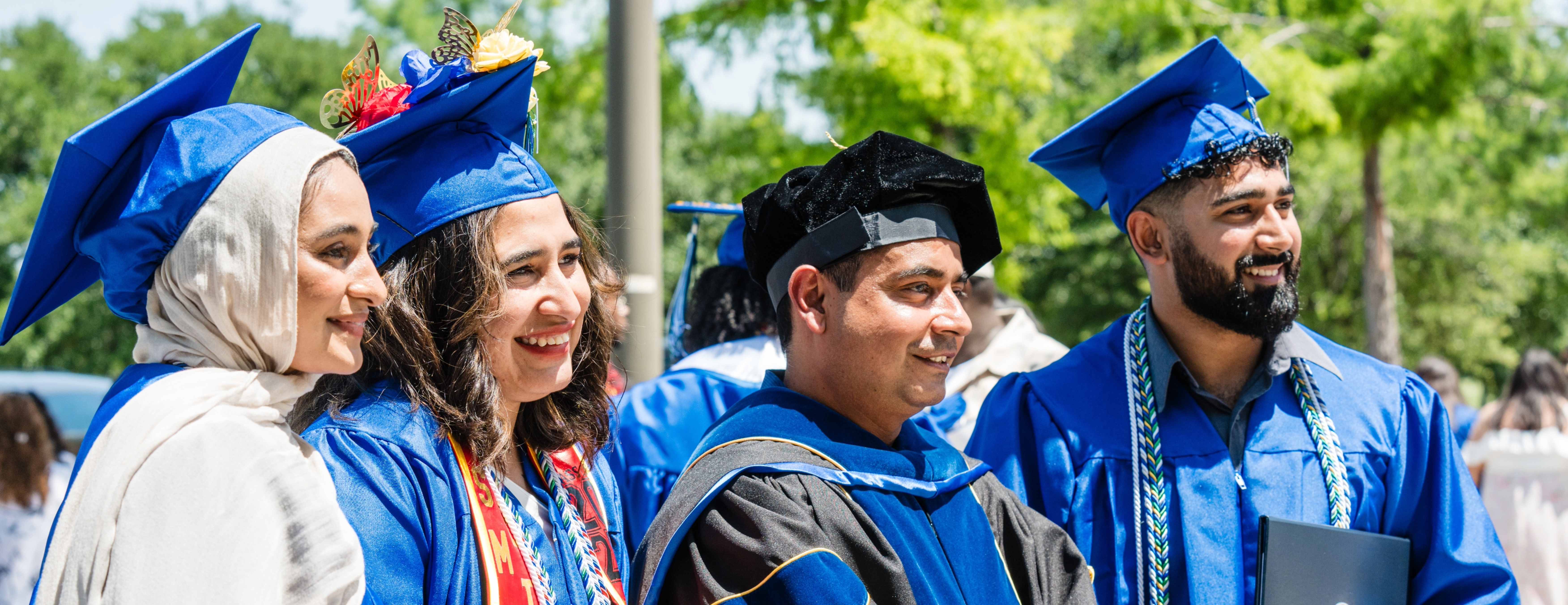 Three graduates and a faculty member smiling for cameras outdoors.