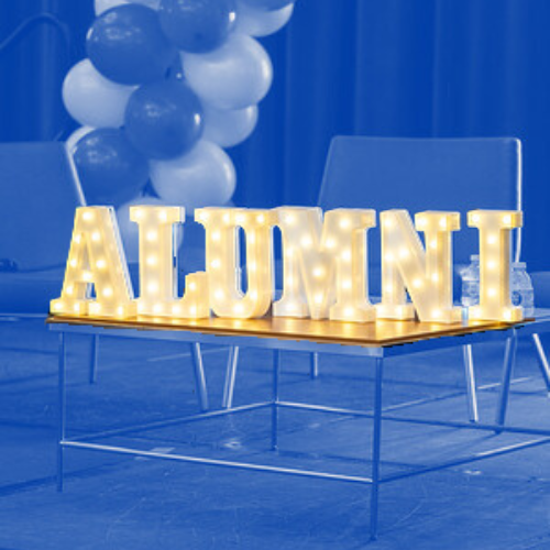 Marquee letters spelling "Alumni" sitting on a table with balloons and chairs in the background.