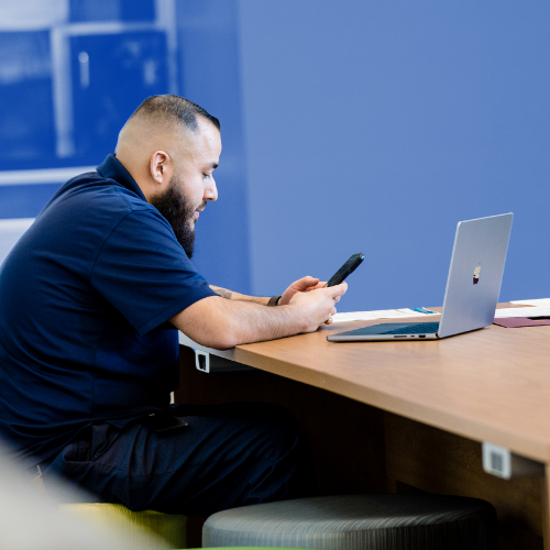 Man sitting at a table and looking down at his mobile device. A laptop is sitting on the table in front of him.