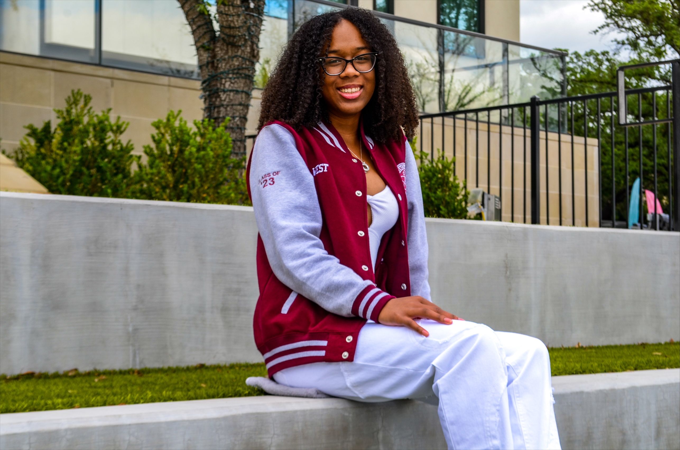 woman with curly hair wearing glasses, a red and gray letterman jacket, and white pants and shirt. She is setting on concrete with a building and foliage in the background