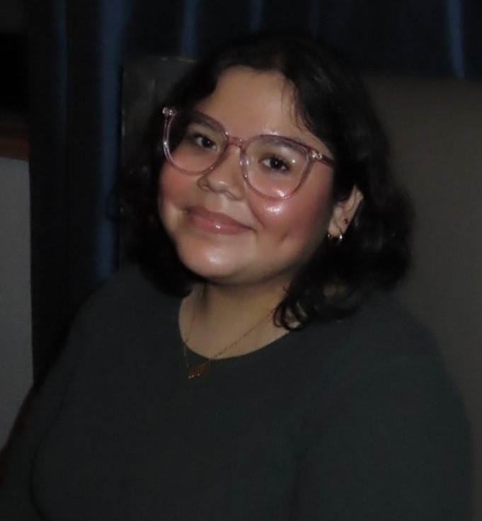 smiling woman in pink glasses and dark blue shirt with dark blue curtains behind her