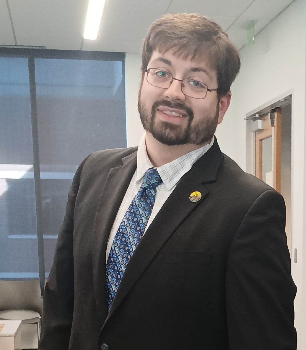 man with short hair and beard smiling, wearing a black suit jacket, blue patterned tie, and a white shirt